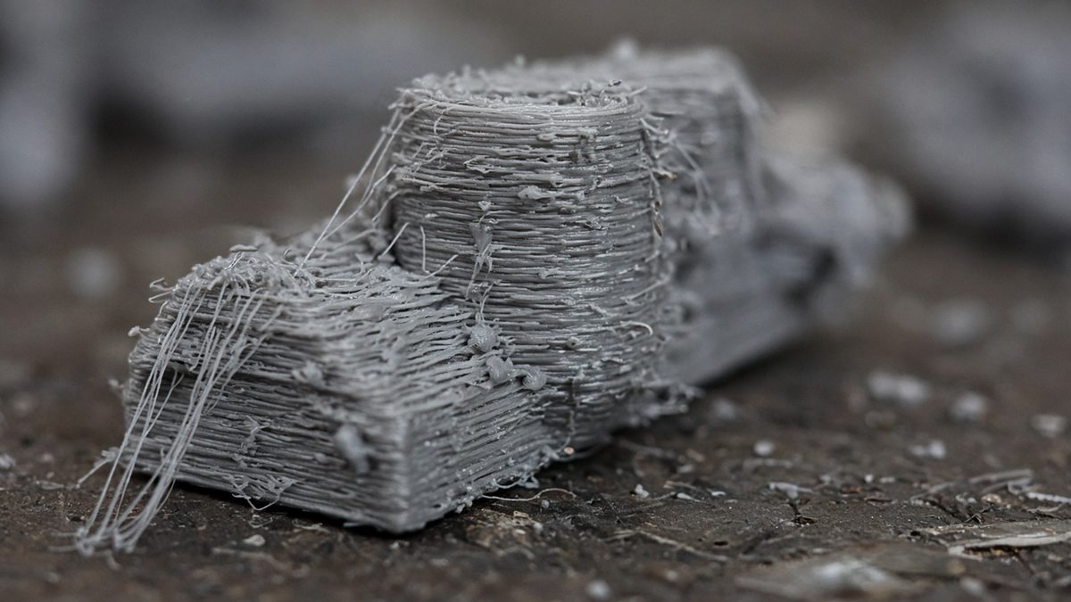 A close-up of a grey 3D print covered in fine stringing hairs, blobs and rough layers caused by wet filament.